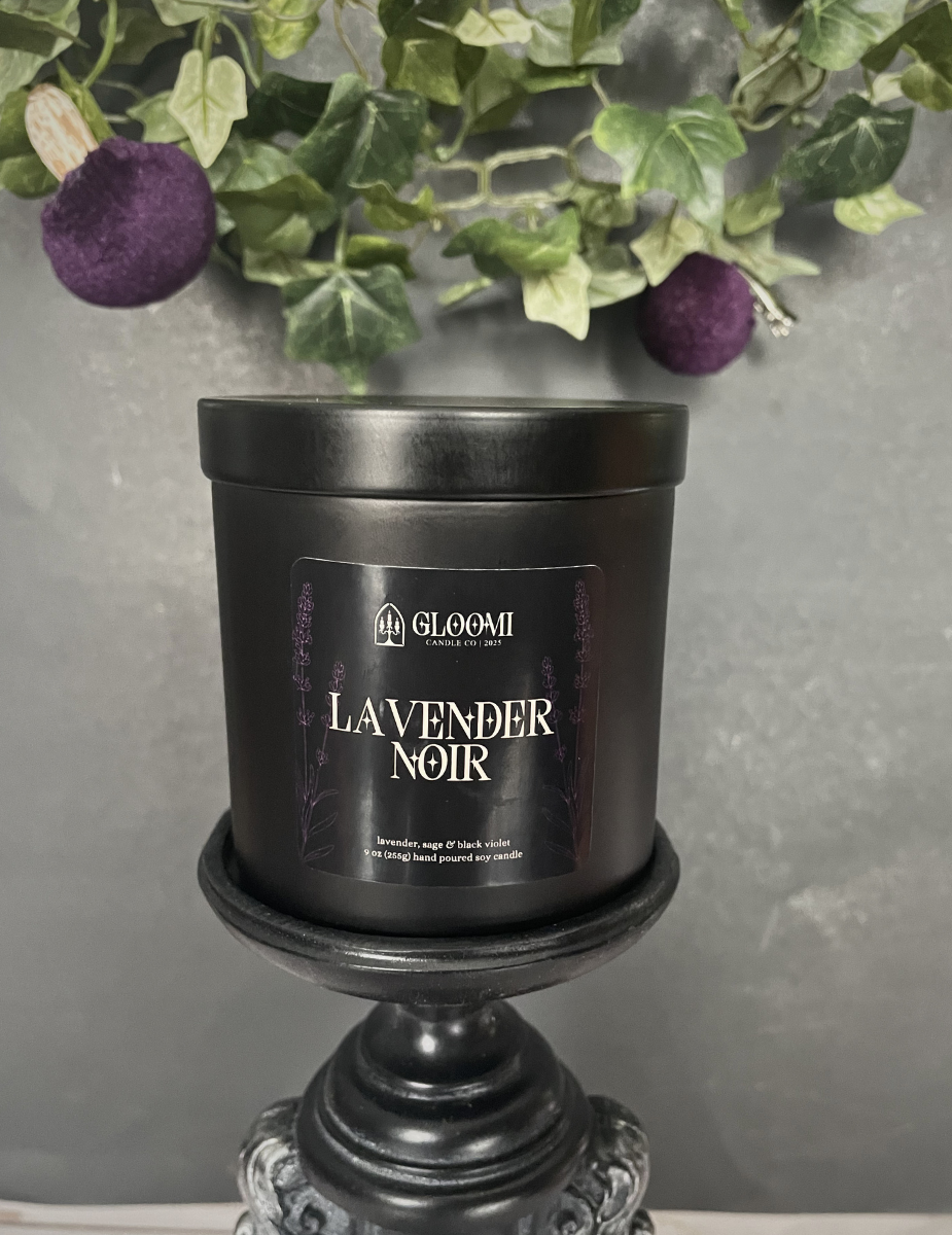Black candle labeled 'Lavender Noir' on a decorative stand with greenery and purple flowers in the background.